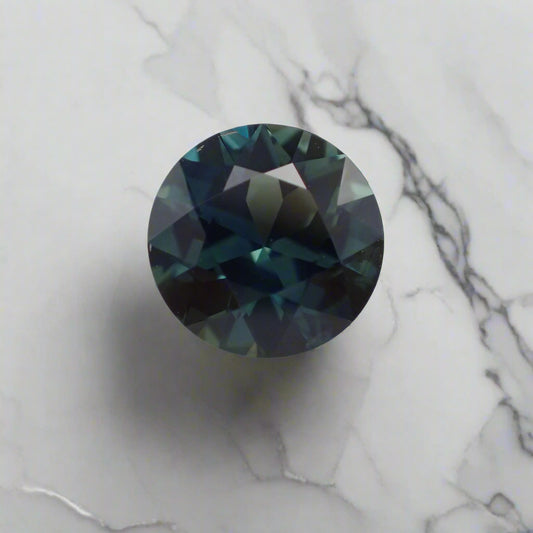 A rich blue and teal sapphire rests on a marble surface. The faceted gem is round.