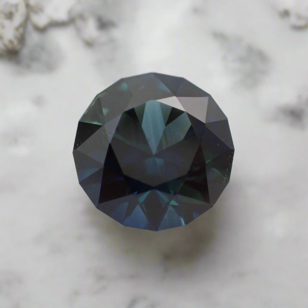 A rich blue sapphire rests on white marble. It is round with a faceted girdle.