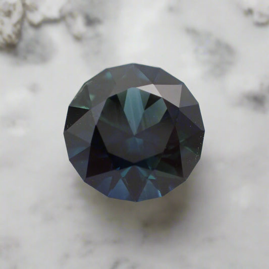 A rich blue sapphire rests on white marble. It is round with a faceted girdle.