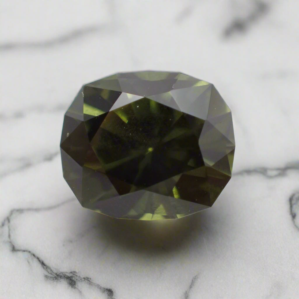 A dark green cushion cut sapphire rests on white marble. The sapphire has a faceted girdle.