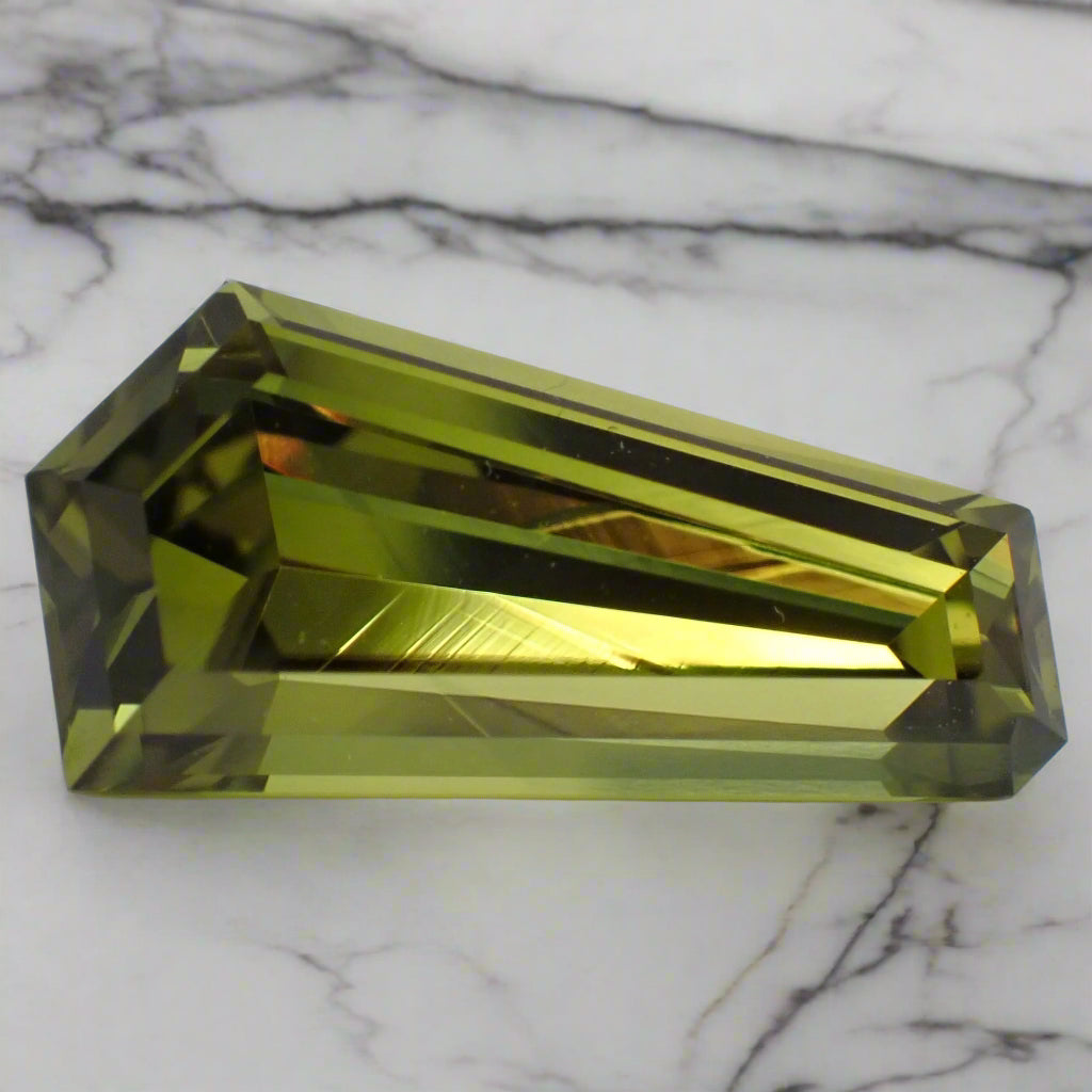 A five sided, freeform sapphire rests on marble. The gem is a bright, olive green.