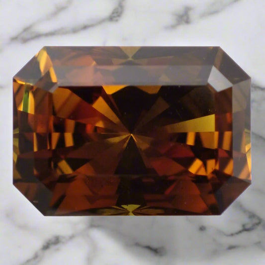 A vivid orange-brown sapphire rests on marble. The sapphire is rectangular with cropped corners.