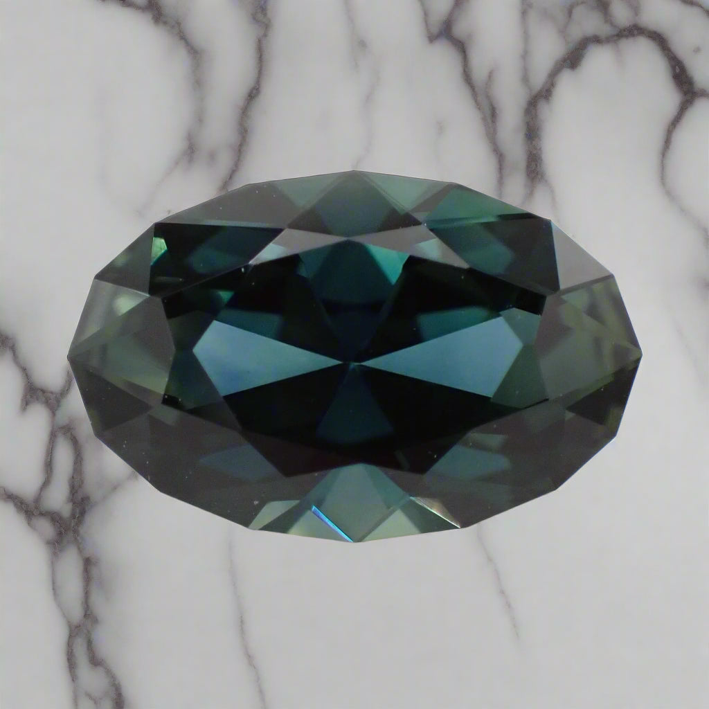 An oval sapphire rests on a white marble suface. The sapphire has a faceted girdle and shows blue, green and teal flashes.