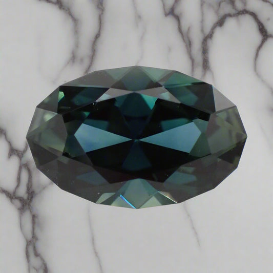 An oval sapphire rests on a white marble suface. The sapphire has a faceted girdle and shows blue, green and teal flashes.