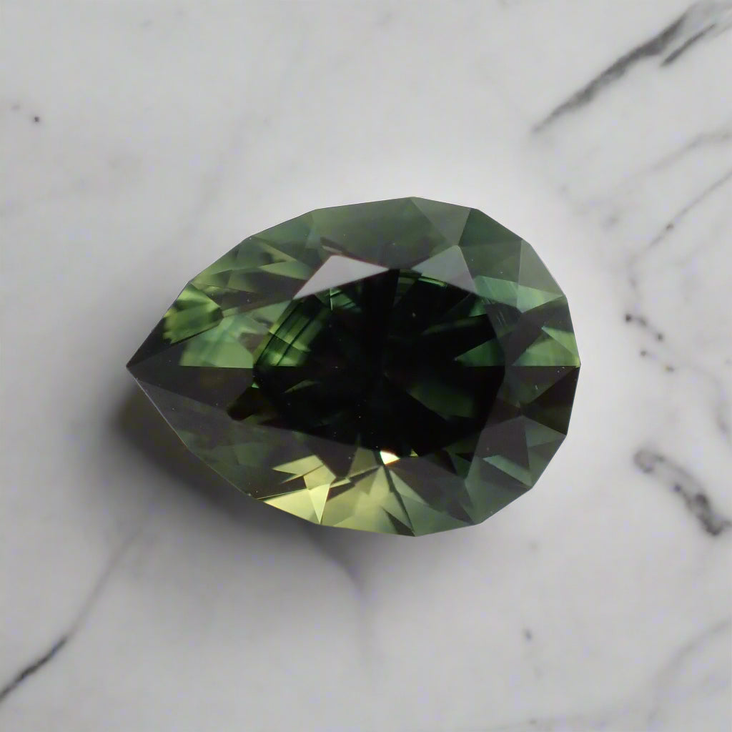 A green, pear cut sapphire with a faceted girdle sits on white marble. There are dark blue colour bands and flashes of yellow visible.