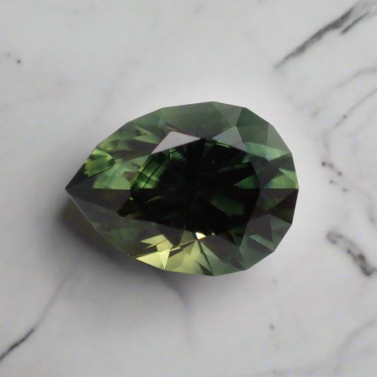 A green, pear cut sapphire with a faceted girdle sits on white marble. There are dark blue colour bands and flashes of yellow visible.