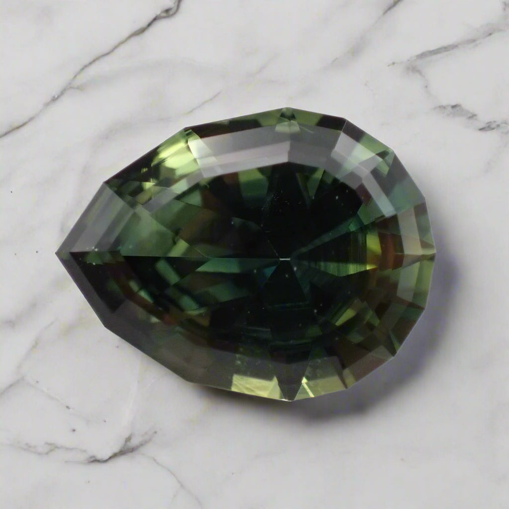 A large, pear shaped sapphire rests on a marble surface. It is predominantly green with some blue and yellow showing.