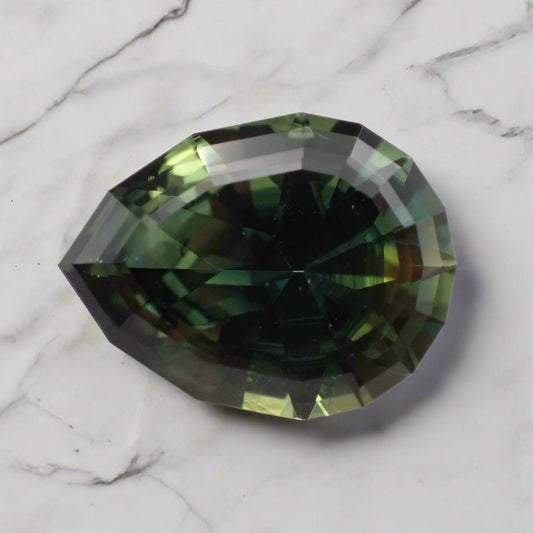 A large, pear shaped sapphire rests on a marble surface. It is predominantly green with some blue and yellow showing.