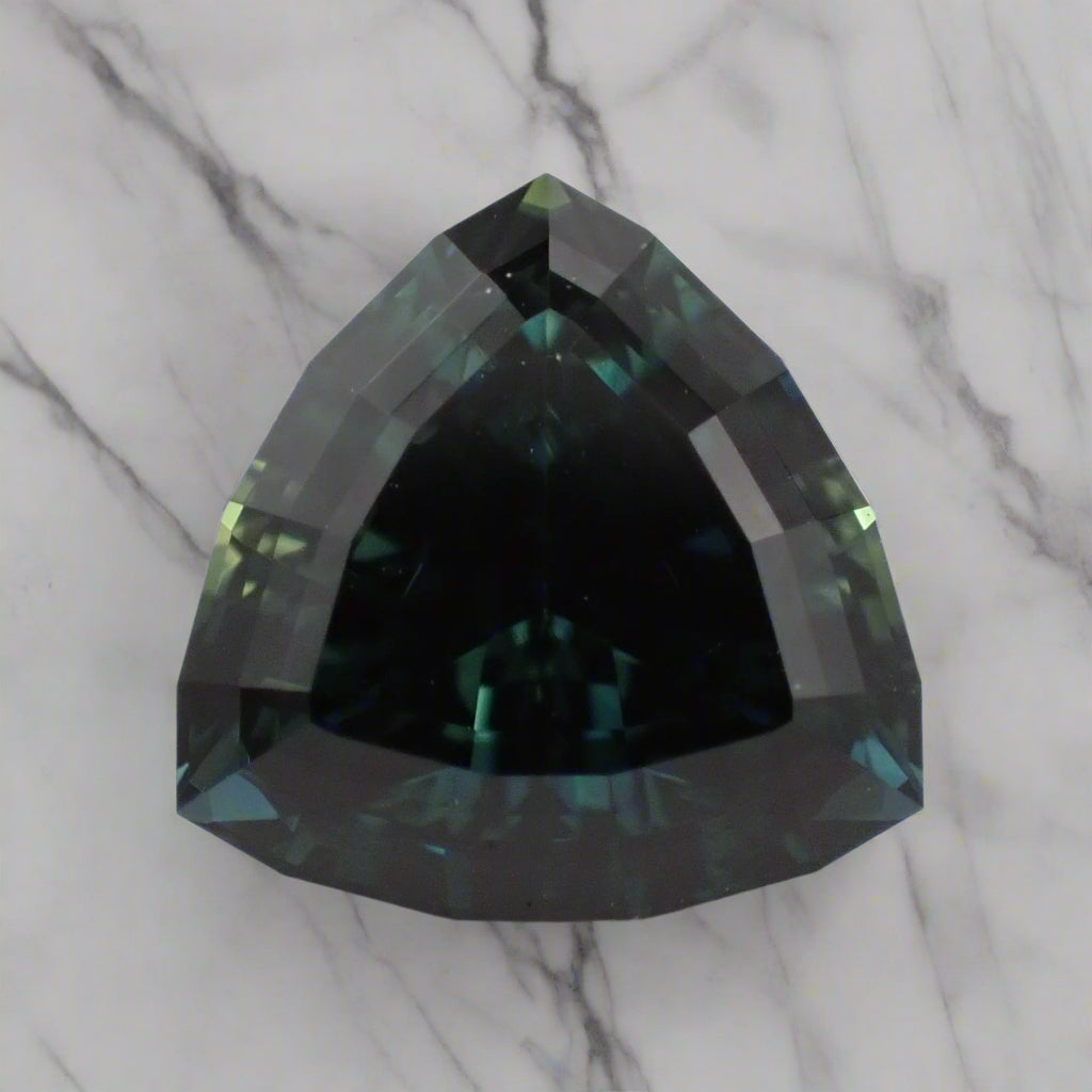 An Australian sapphire, showing blue, green and teal, rests on a marble surface. The sapphire is a triangle shape with a faceted girdle.