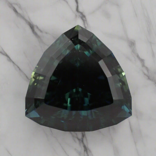 An Australian sapphire, showing blue, green and teal, rests on a marble surface. The sapphire is a triangle shape with a faceted girdle.