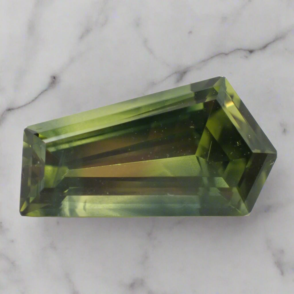 A mainly green, with patches of blue, sapphire rests on marble. The gem is freeform in shape with five sides.