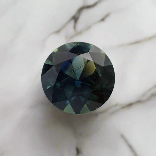 A predominantly blue sapphire rests on white marble. The sapphire shows flashes of royal blue, light teal and yellow.