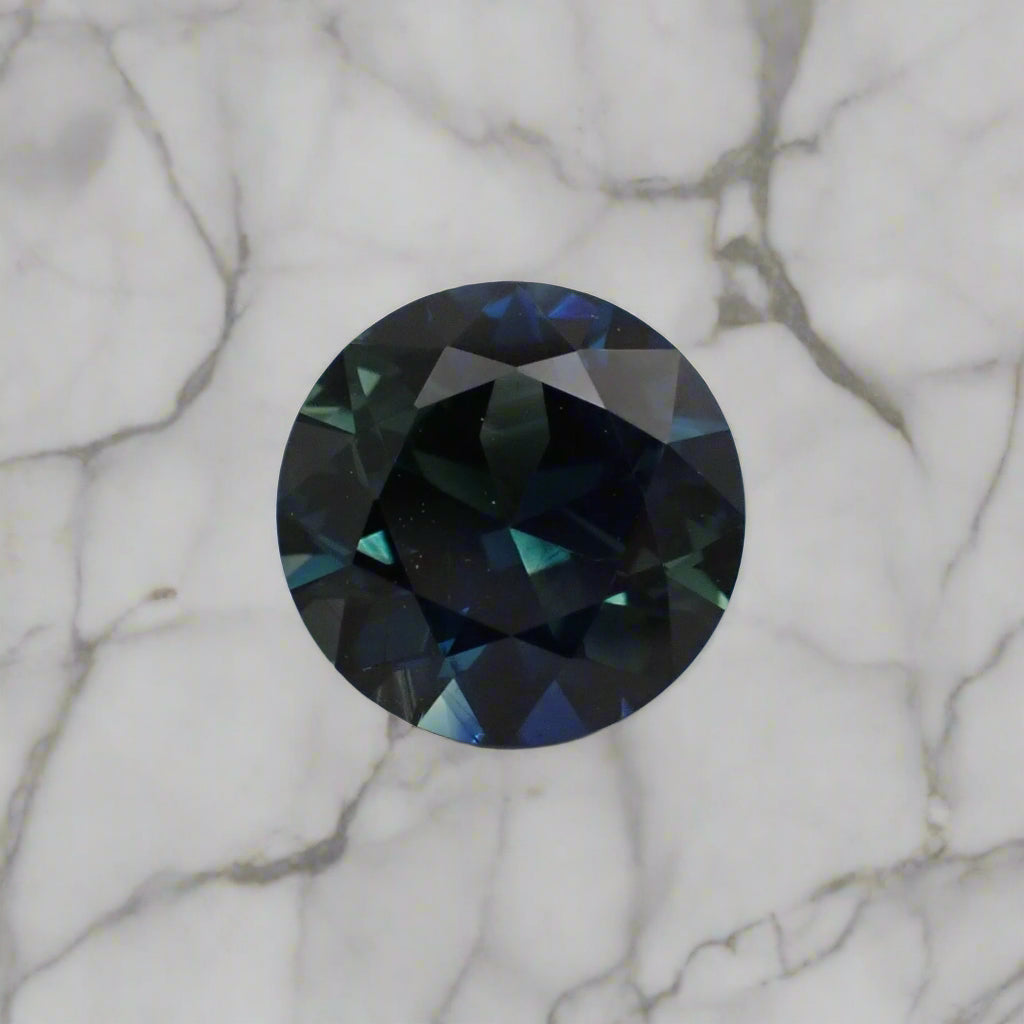 A bright round sapphire with marble out of focus in the background. The sapphire is a mix of blue and green.