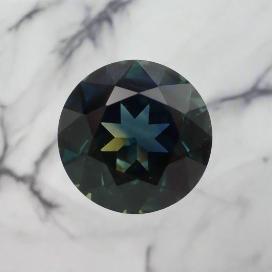 A round sapphire with a mix of blue and yellow colour banding combining in places to make green. The gem is resting on a marble surface.