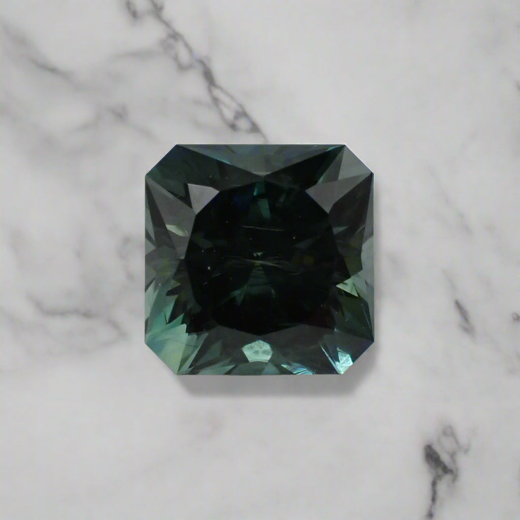 A square sapphire with cropped corners rests on a marble surface. The sapphire shows flashes of light blue and green.