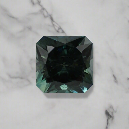 A square sapphire with cropped corners rests on a marble surface. The sapphire shows flashes of light blue and green.