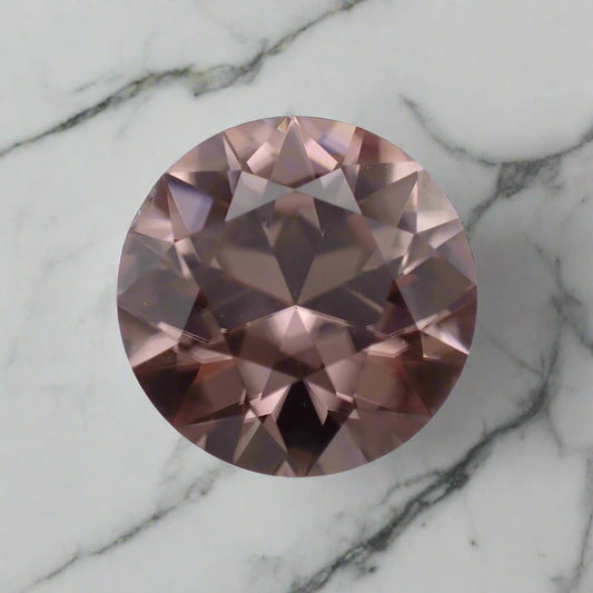 A dusty pink zircon rests on a marble surface. The gem is round, clear and bright.