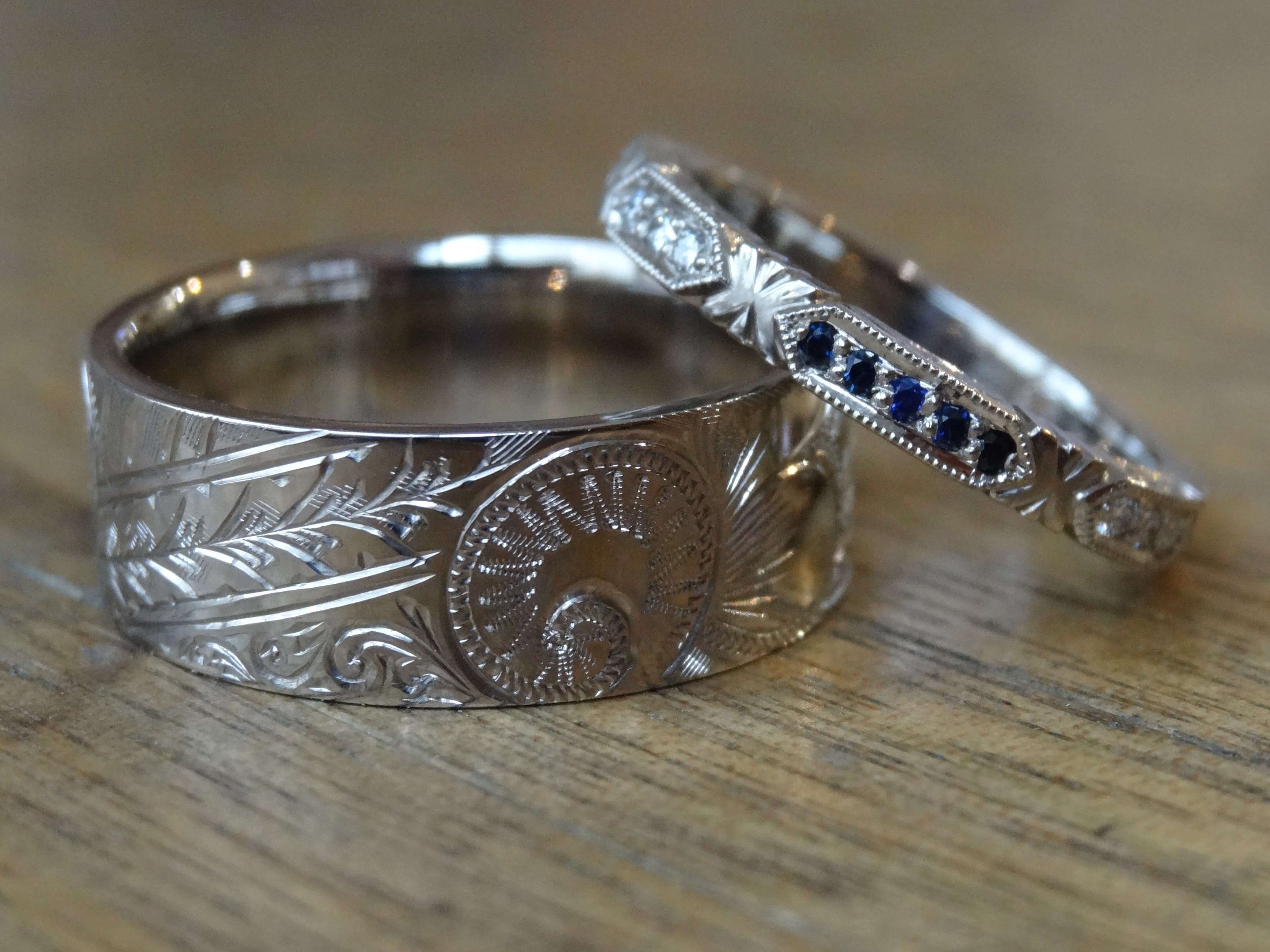 Wide palladium hand engraved gents wedding sitting below a narrow ladies wedding ring that is hand engraved and set with small blue sapphires
