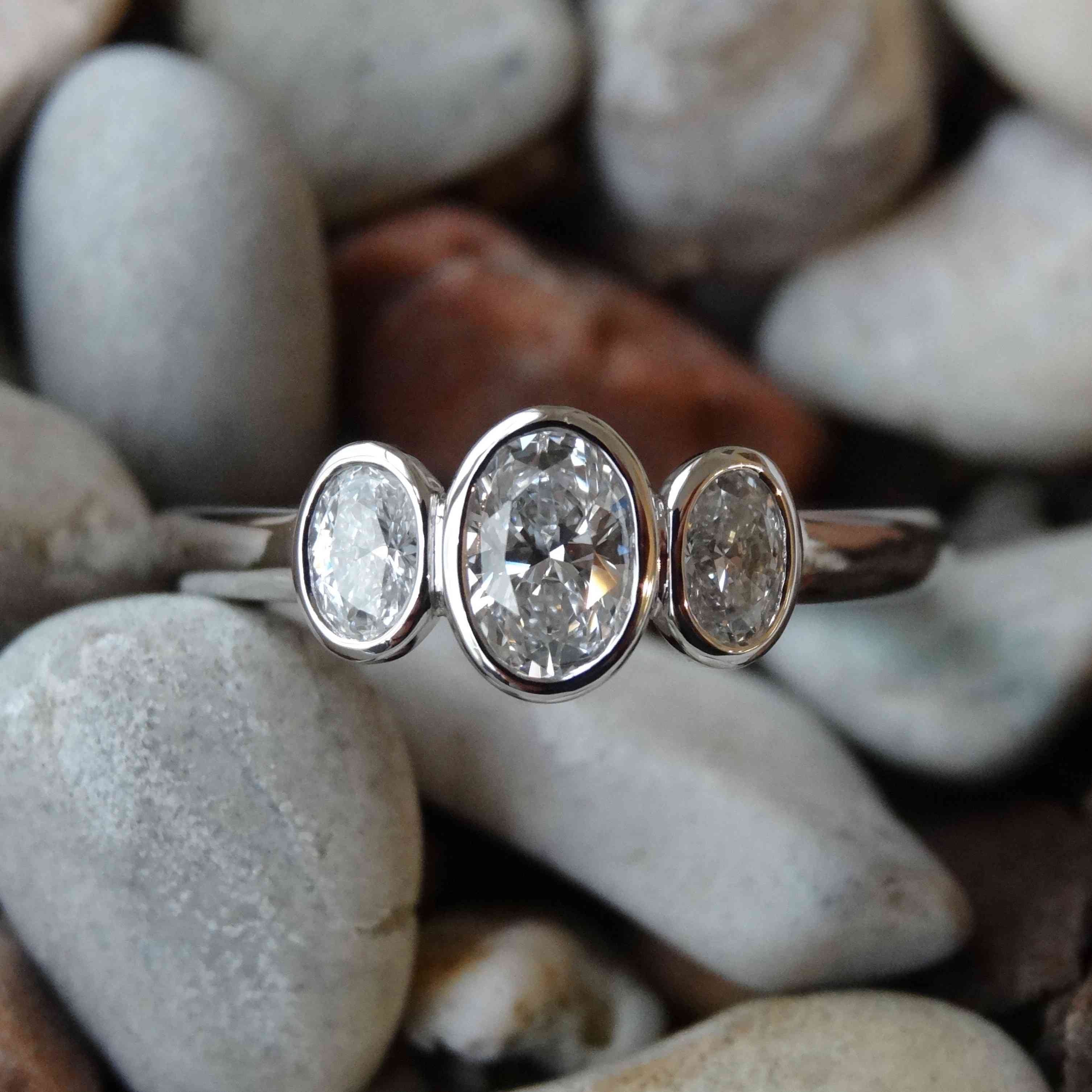 Platinum trilogy engagement ring bezel set with three oval diamonds laying on small grey pebbles