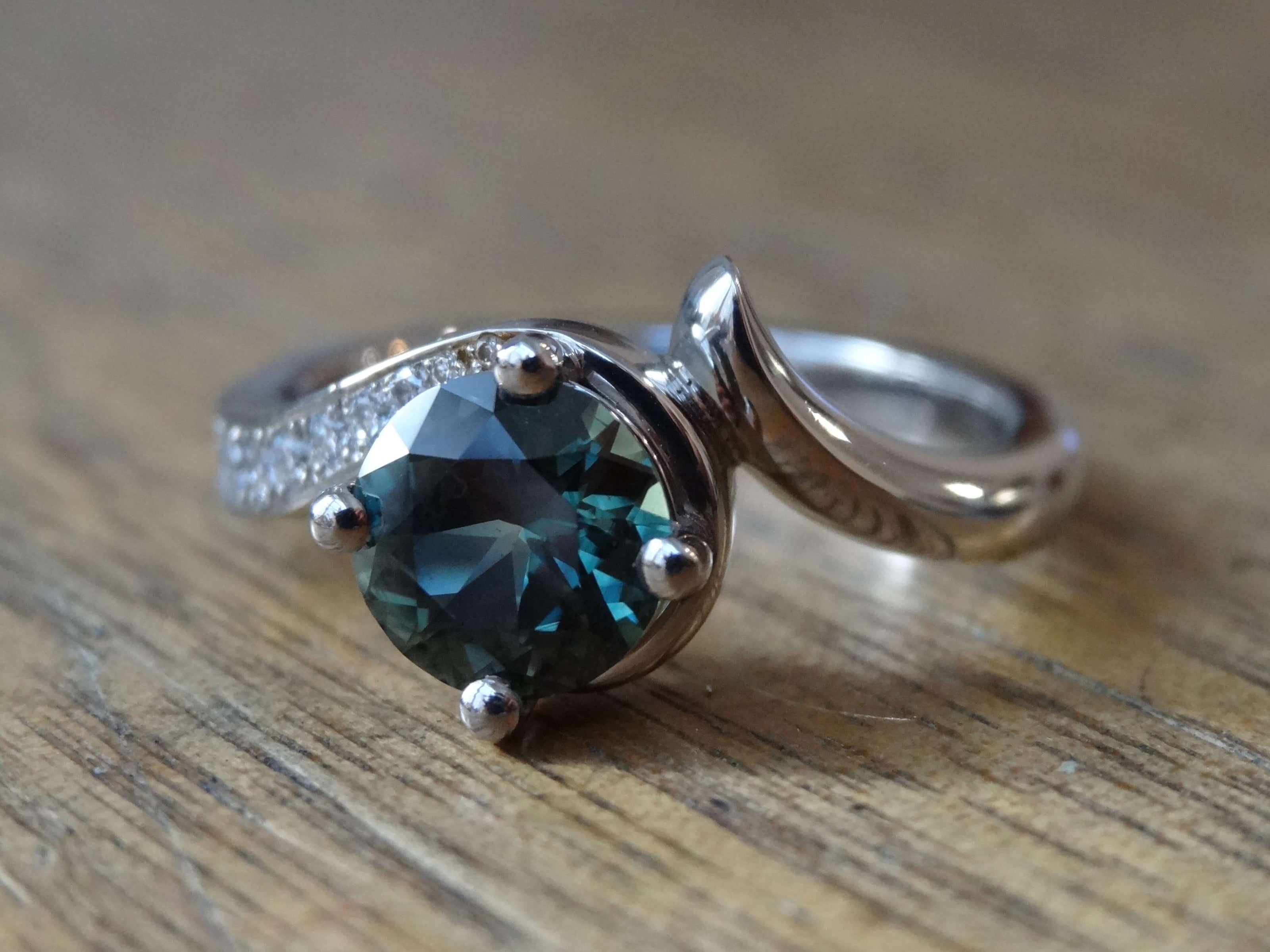 Custom made palladium swirl engagement ring claw set with one round teal sapphire and small diamonds grain set in the band.