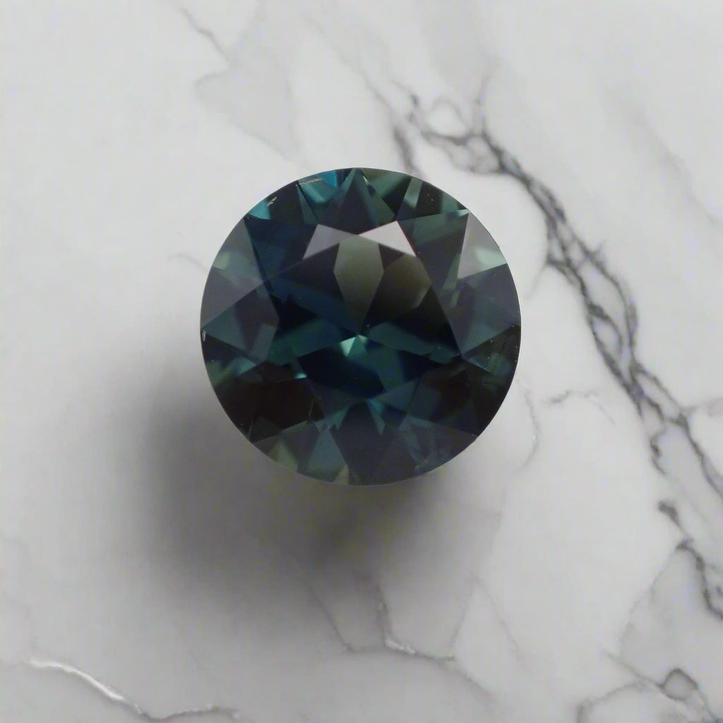 A rich blue and teal sapphire rests on a marble surface. The faceted gem is round.
