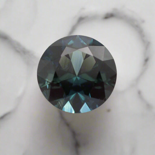 A round blue sapphire rests on white marble. The sapphire shows flashes of light blue, teal and denim colours.