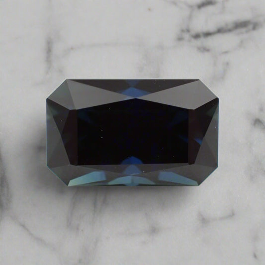 A dark, rich blue sapphire, rectangular in shape, rests on a white marble surface