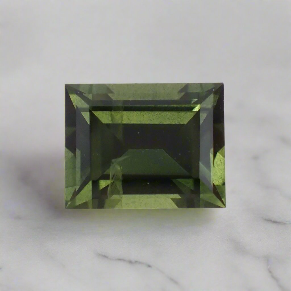 A forest green sapphire, rectangular baguette cut, rests on white marble.