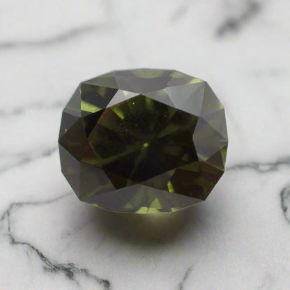 A dark green cushion cut sapphire rests on white marble. The sapphire has a faceted girdle. 