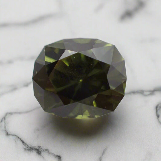 A dark green cushion cut sapphire rests on white marble. The sapphire has a faceted girdle. 