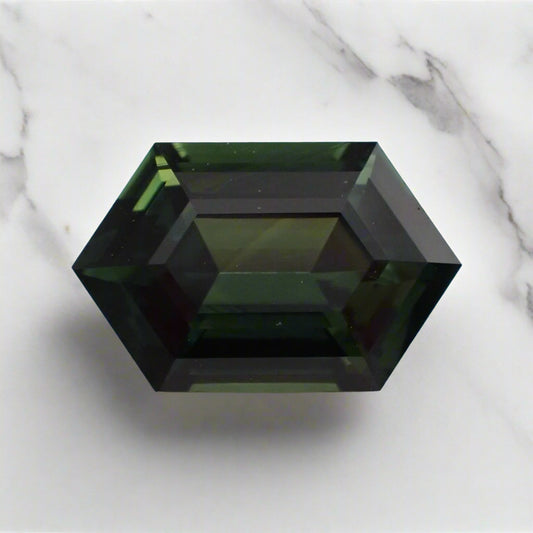 A dark forest green sapphire, elongated hexagon in shape, rests on a white marble surface.