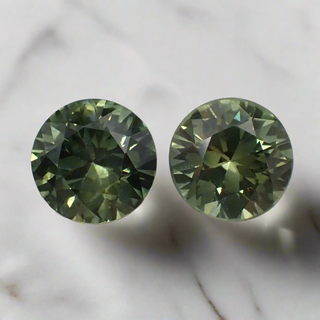 a pair of round, bright, mid green sapphires rest on a white, marble surface