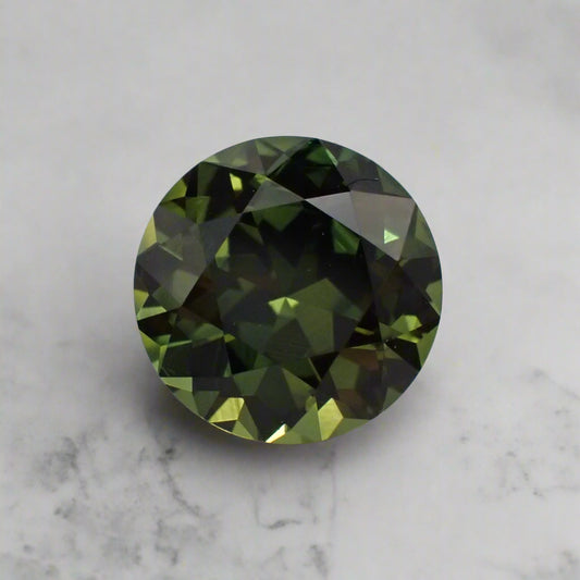 An olive green sapphire, with lots of flashes of colour, rests on a white marble surface.