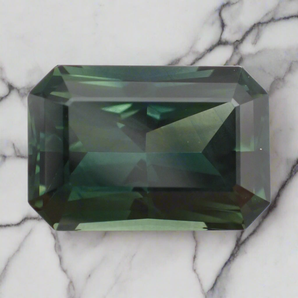 A rich, forest green sapphire rests on marble. The radiant cut stone is rectangular with cropped corners.