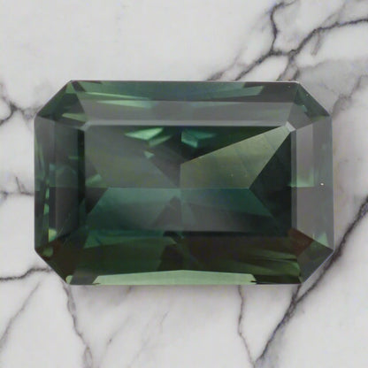 A rich, forest green sapphire rests on marble. The radiant cut stone is rectangular with cropped corners.