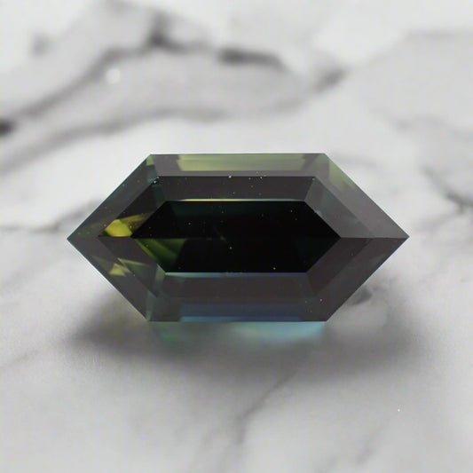 An elongated hexagonal sapphire rests on a white surface. The top left corner of the stone shows lots of yellow and the bottom edge mainly blue.