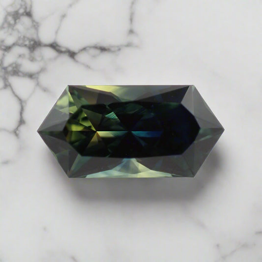 A loose sapphire rests on a white marble background. It is an elongated hexagon shape and shows bright flashes of green, blue and yellow.