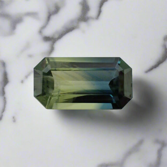 A parti sapphire, with distinct blue/yellow division and visible silt, rests on a white marble surface.