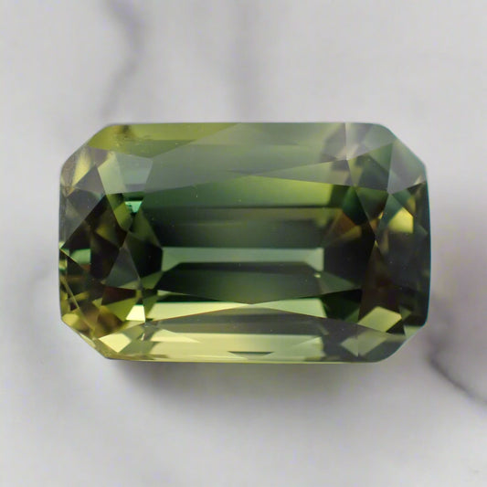A light, bright sapphire rests on white marble. The stone is a mix of green and yellow. 