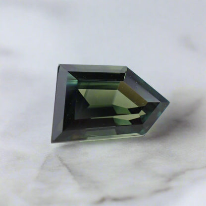 A dark sapphire showing blues and greens, cut in a five sided freeform shape, rests on a white surface.