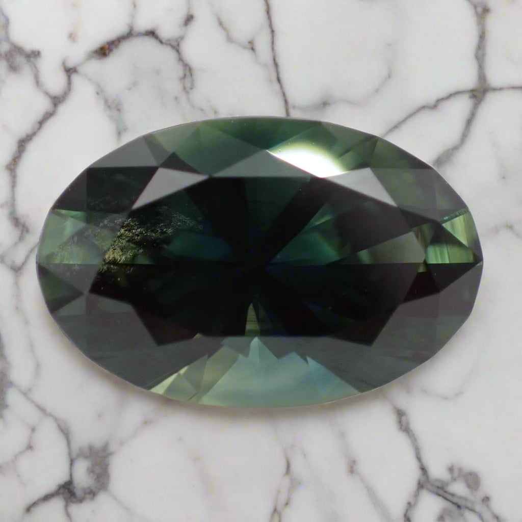 A large oval sapphire rests on marble. The sapphire shows greens, blues and teal flashes and some inclusions can be seen on the left side. 