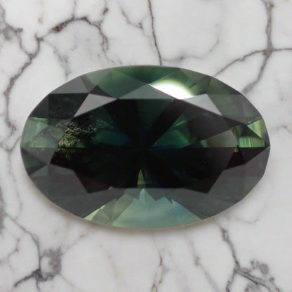 A large oval sapphire rests on marble. The sapphire shows greens, blues and teal flashes and some inclusions can be seen on the left side. 