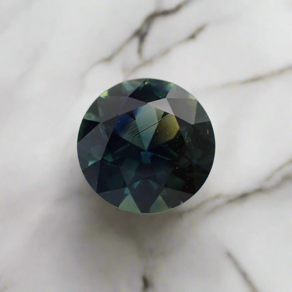 A predominantly blue sapphire rests on white marble. The sapphire shows flashes of royal blue, light teal and yellow.