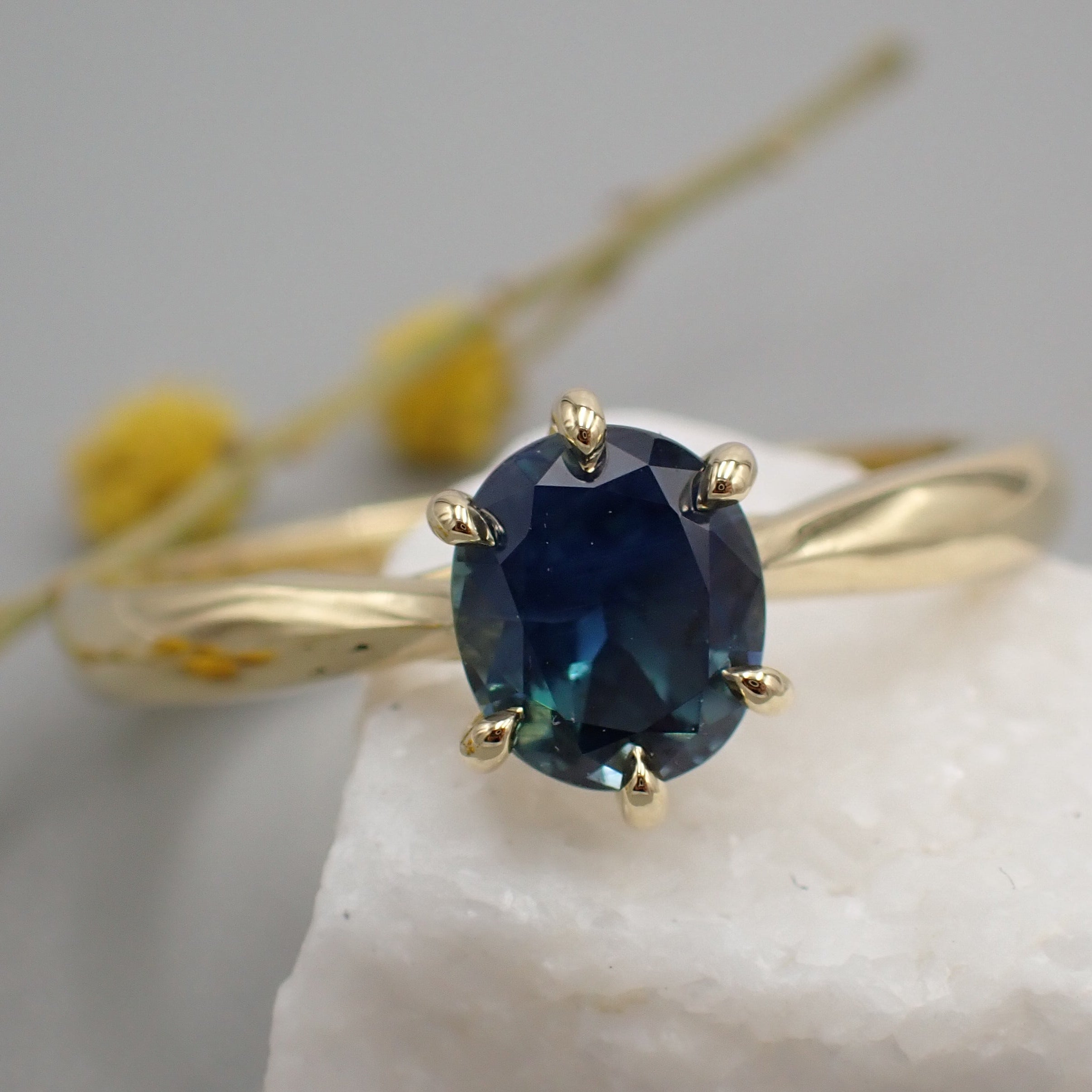 A blue sapphire with flashes of green and yellow is set in a six claw, yellow gold solitaire engagement ring. The ring rests on a white rock with some yellow flowers out of focus in the background.