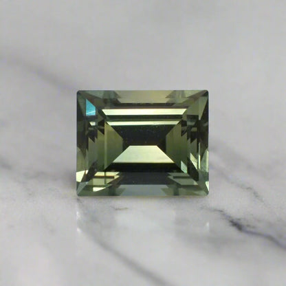 A light teal, baguette cut sapphire rests on a white marble surface.