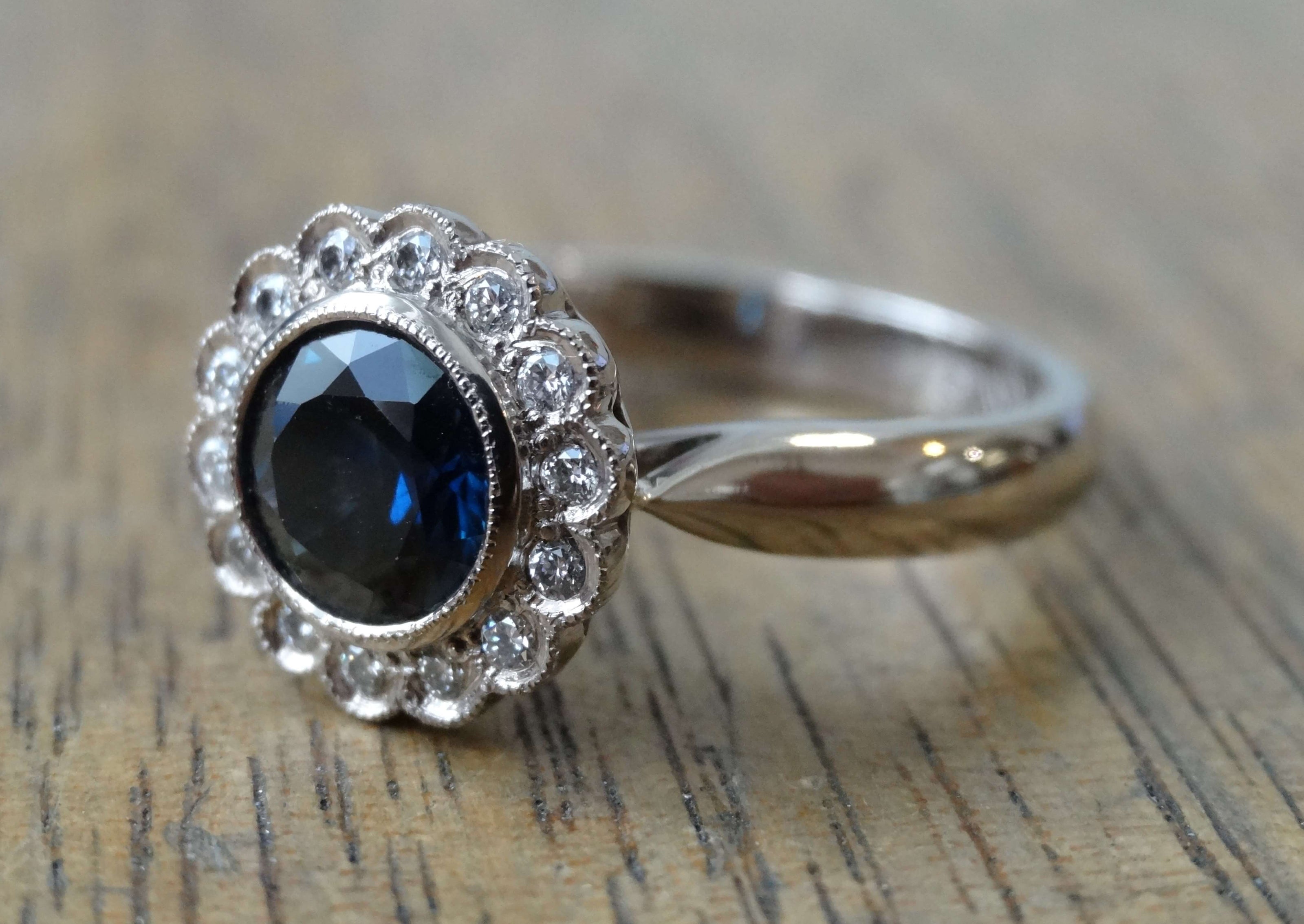 Platinum engagement ring set with a round blue sapphire and a halo of diamonds with mill-grain.  Ring is lying on a wooden surface