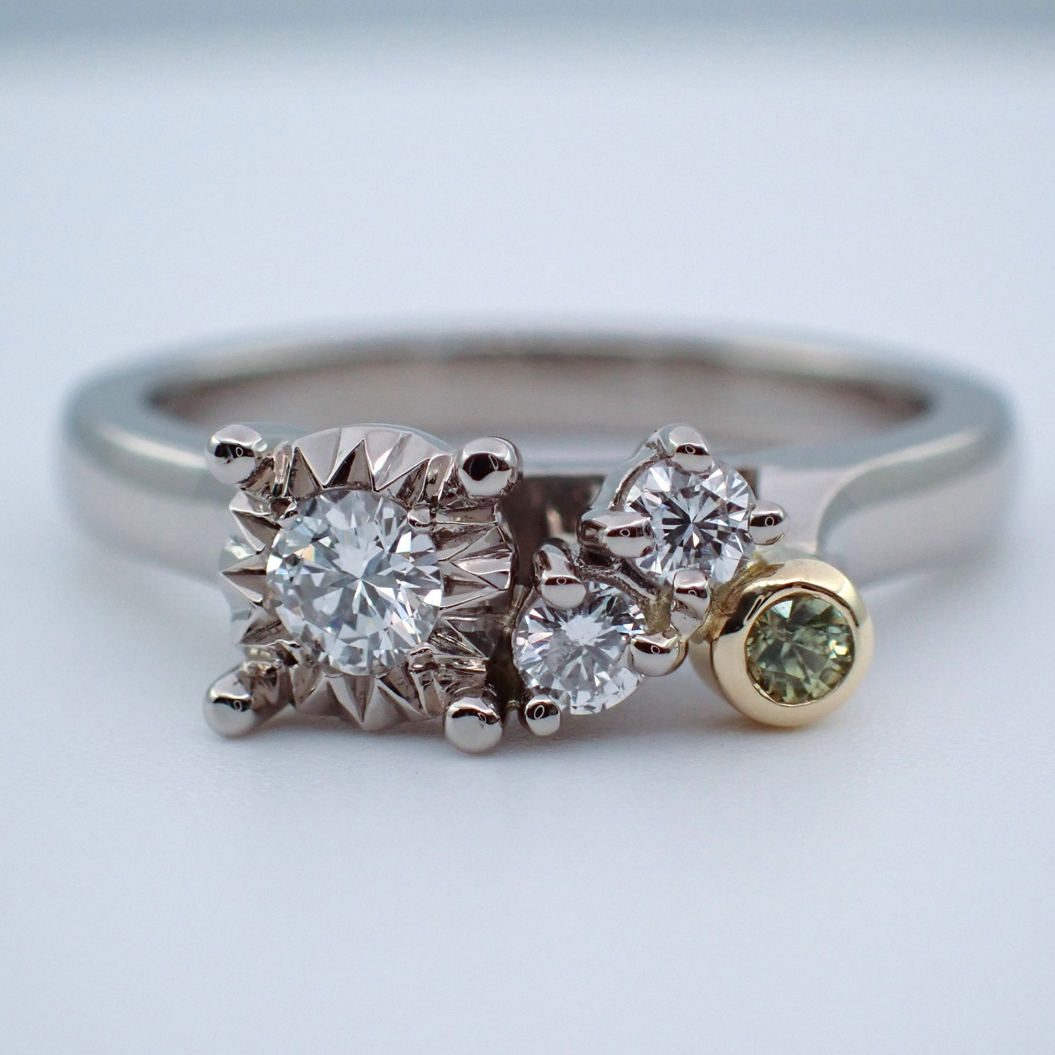 An engagement ring set with three round diamonds and one green sapphire. The ring is both white and yellow gold with a mixture of setting styles.