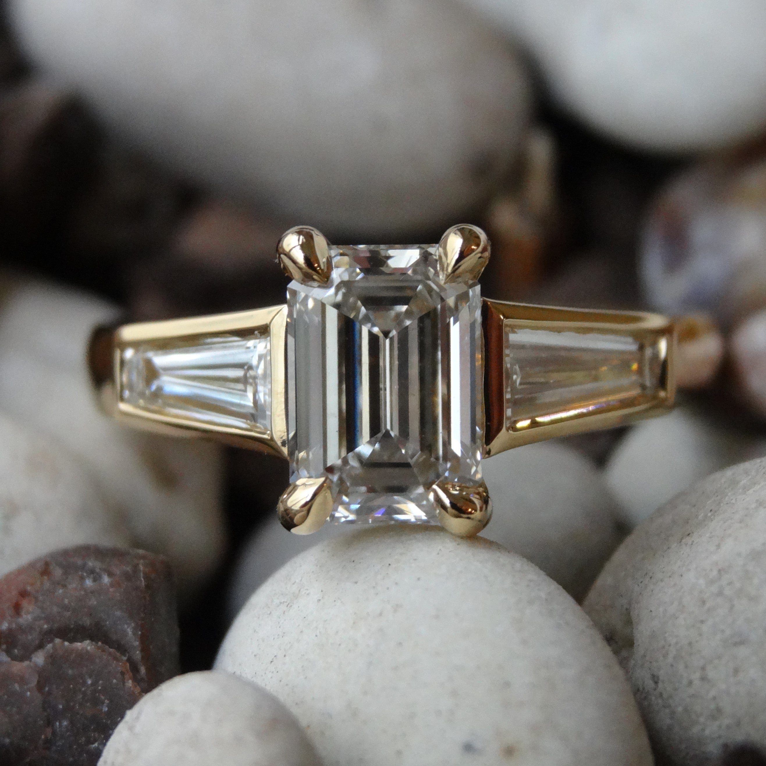 A three stone engagement ring rests on decorative rocks. The emerald cut diamond is set in claws with tapered baguettes bezel set on either side.