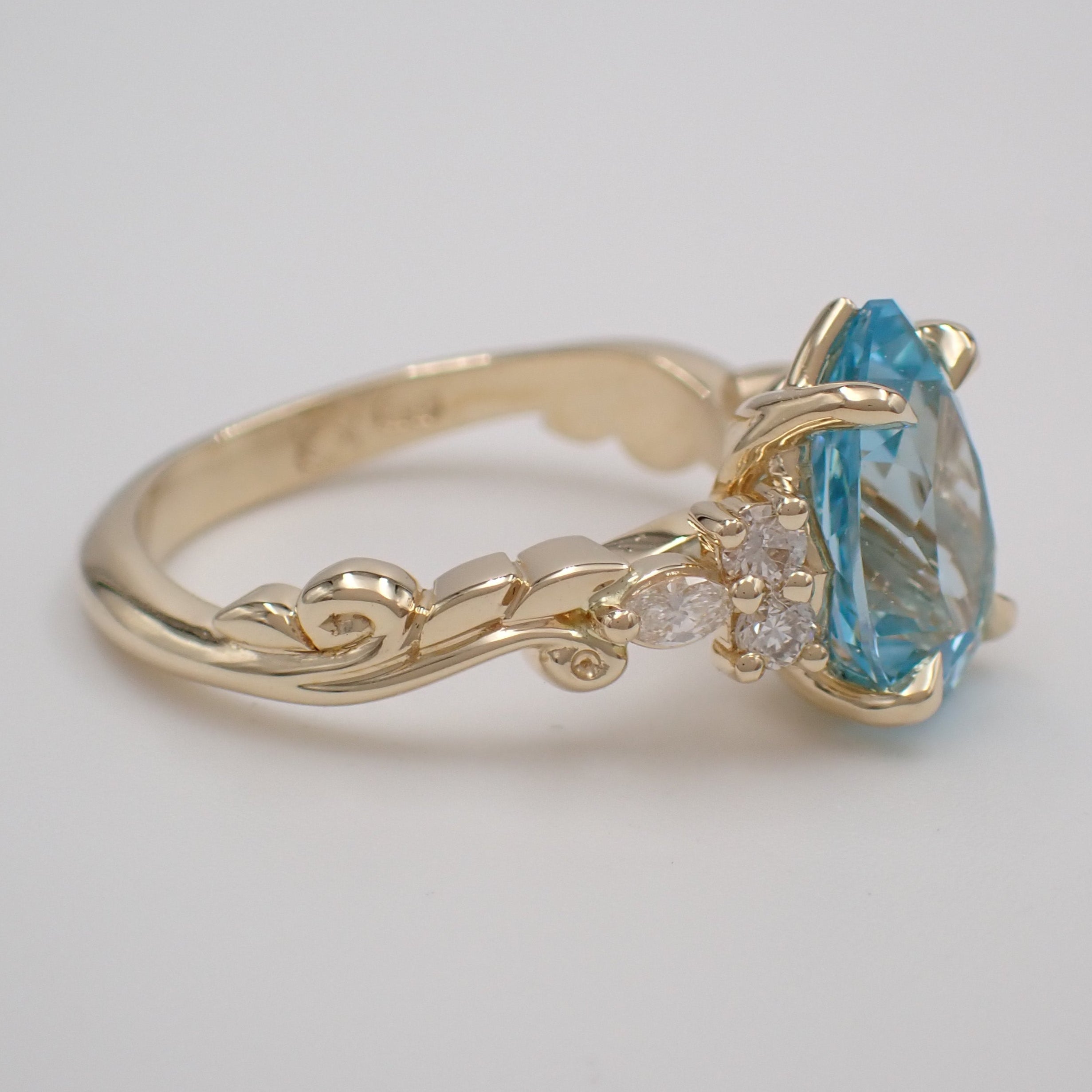 A pear cut blue sapphire is claw set with three small lab grown diamonds set on either side. The band of the ring is a carved vine and leaf design.