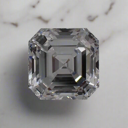 A loose, faceted diamond rests on a white background. It is a square emerald cut stone.